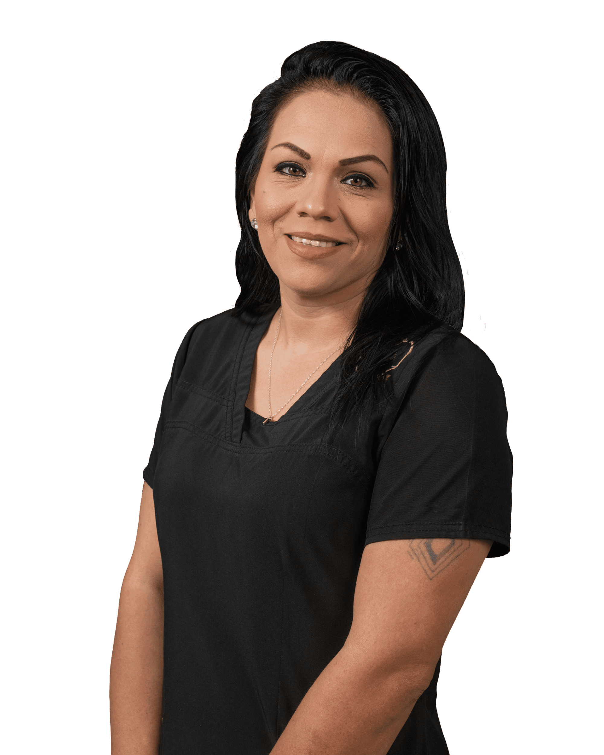 A woman with long dark hair, wearing a black short-sleeve top, stands smiling with her arms relaxed at her sides. She has a tattoo on her left upper arm.