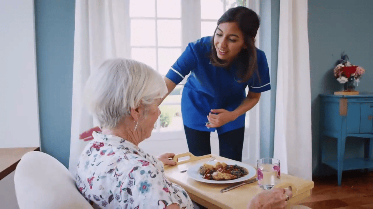 A caregiver in blue scrubs smiles and talks to an elderly woman who is sitting at a table with a meal and a glass of water.
