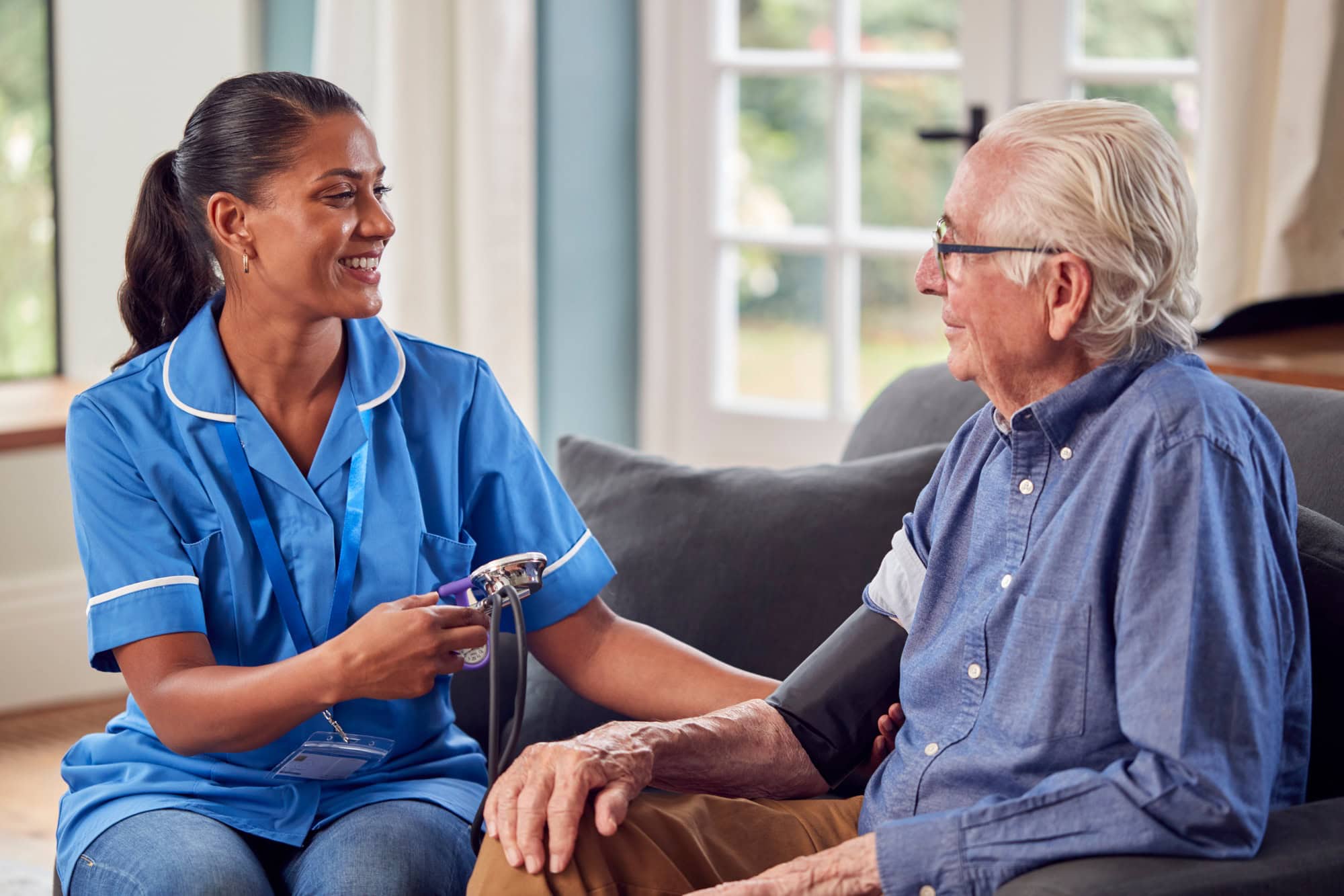 Senior Man At Home In Lounge Having Blood Pressure Taken By Female Care Worker In Uniform