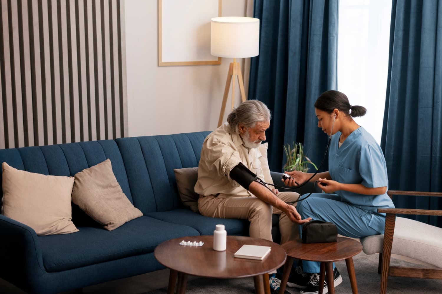 A nurse in blue scrubs measures an elderly man's blood pressure in a living room, setting up a safe recovery space after hospital discharge. Medicine bottles and equipment are visible on the table in front of them.