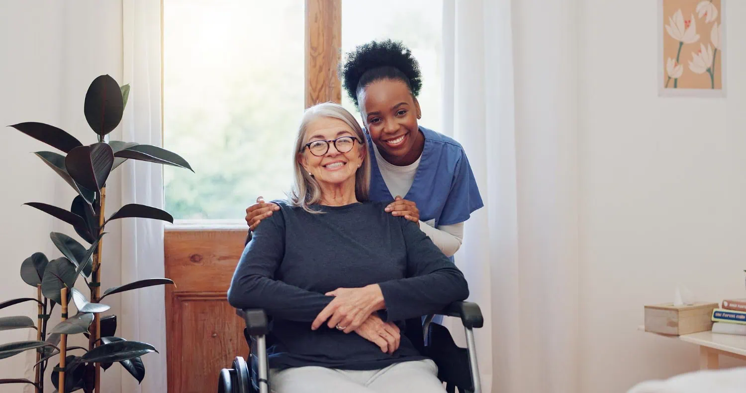An elderly woman in a wheelchair sits smiling, while a caregiver from a team of home care providers stands behind her with hands on her shoulders, both smiling in a well-lit room.