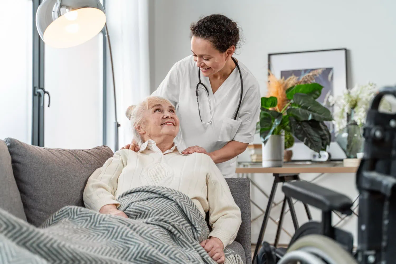 A nurse providing in-home care stands behind an elderly woman sitting on a couch, smiling at her. The woman is covered with a blanket, and a wheelchair is visible in the foreground.