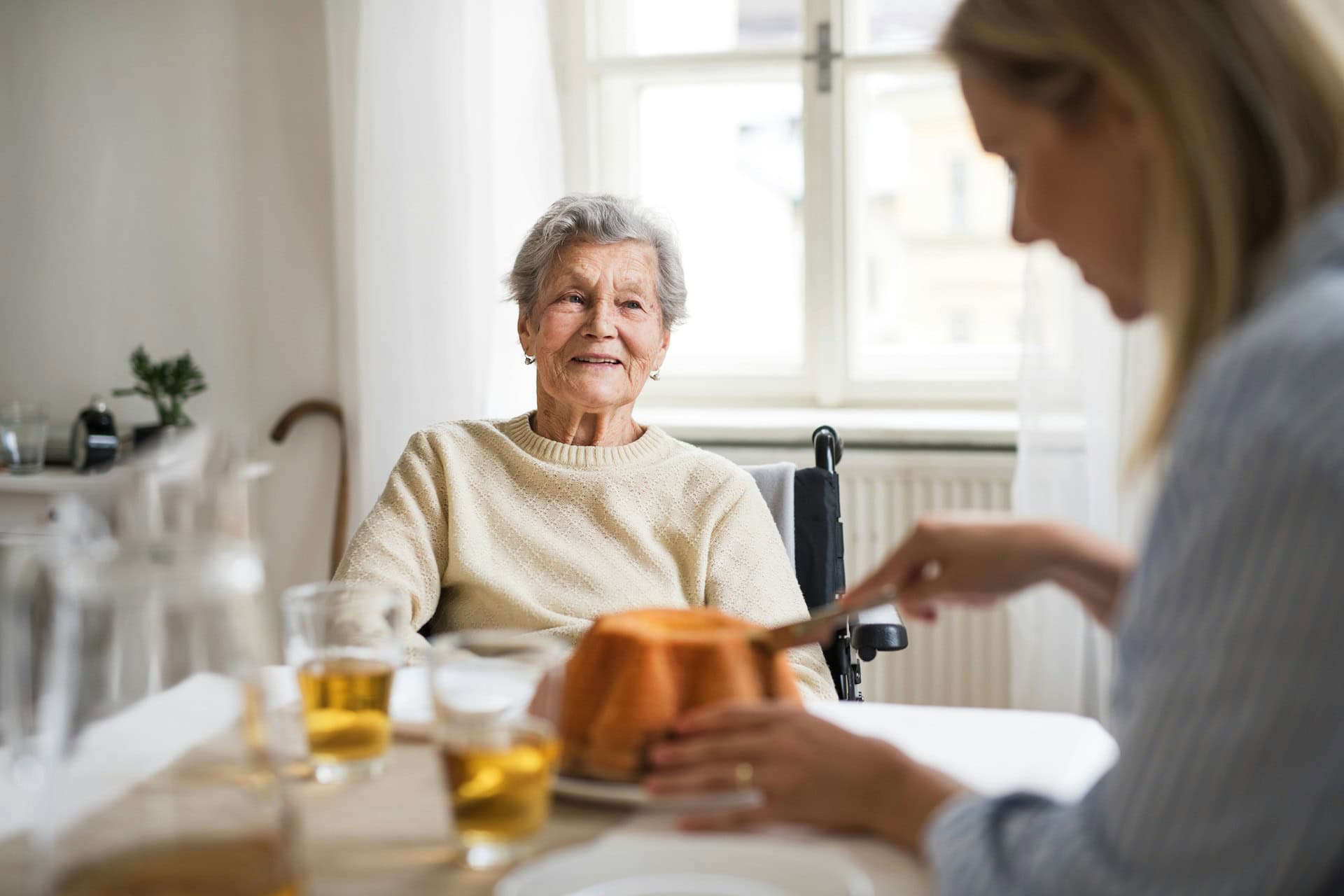 An older woman in a wheelchair sits at a table, smiling, while a younger woman slices a cake in a bright, sunlit room—celebrating the holiday seasons with home care services in Brazoria County.