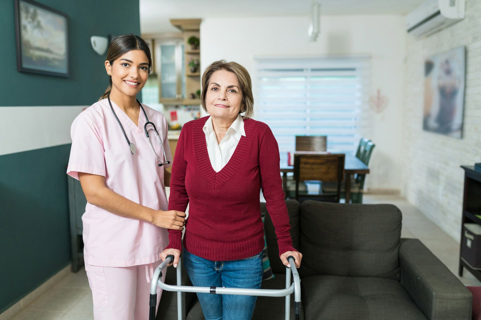 A nurse in pink scrubs stands beside an older woman using a walker in a living room, both smiling at the camera—capturing a warm moment of senior care and support.
