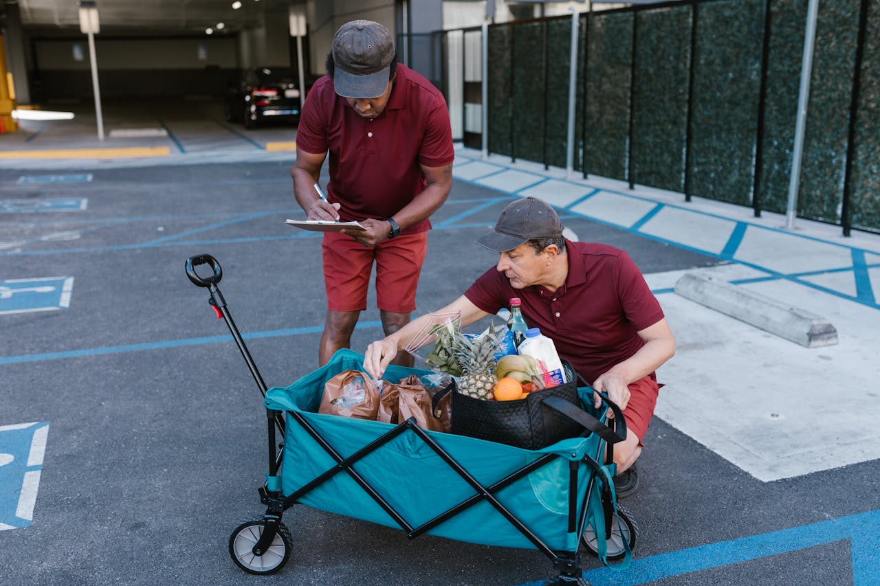 Two men in red shirts and shorts, one writing on a clipboard and the other organizing groceries in a blue wagon, are in a parking lot with marked handicapped spaces.