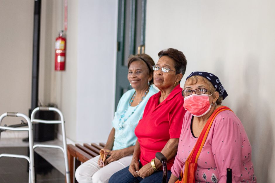 Three elderly women sit on a bench indoors; one wears a face mask and headscarf, and a walking aid is visible beside them, highlighting the warm community atmosphere found in affordable senior care settings throughout Brazoria County.
