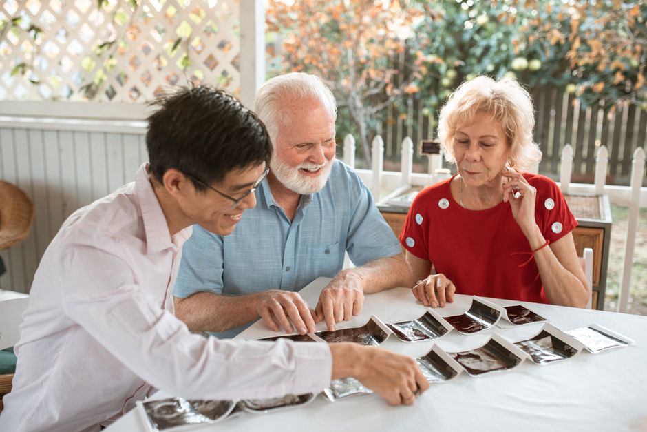 Three people sit at a table outdoors, looking at several black and white photos arranged in rows, perhaps reminiscing as they explore memories together—an intimate moment that reflects the importance of connection highlighted in any good senior care guide.