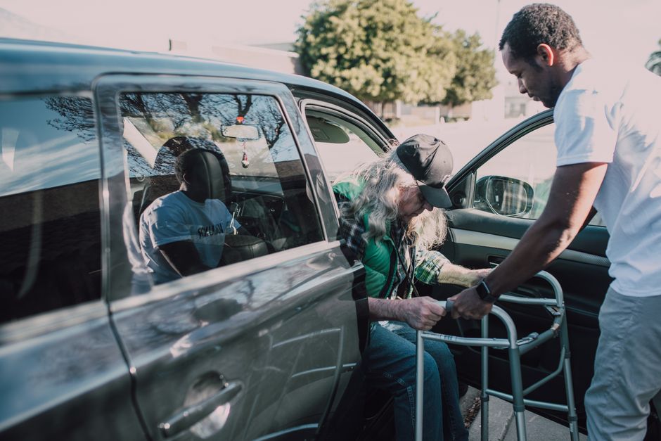 An older man with a walker exits a car with the assistance of another man standing nearby on a sunny day, highlighting the importance of transportation assistance for seniors in Pearland.