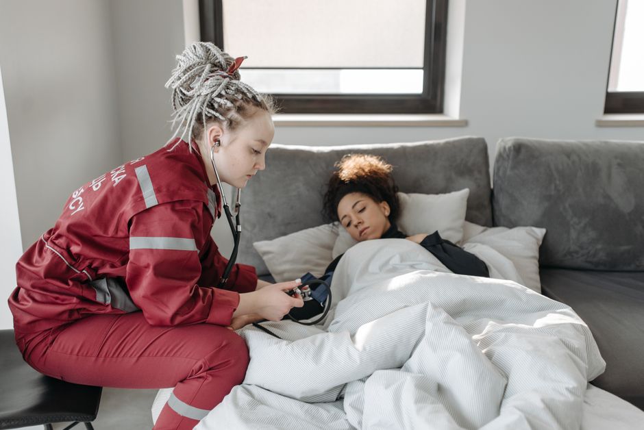A paramedic in a red uniform checks the blood pressure of a woman lying in bed, illustrating attentive recovery at home in a well-lit room.