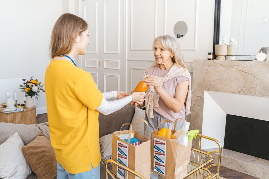 A younger woman hands a bottle to an older woman in a living room, with grocery bags full of food on a cart between them—a warm moment that highlights the importance of caregiver support in daily life.