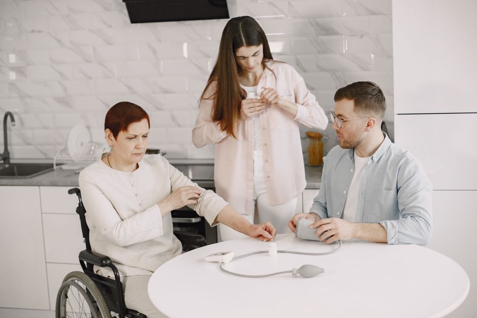 A woman in a wheelchair has her blood pressure checked by a man at a table while another woman—likely one of her caregivers—stands nearby in a kitchen setting.