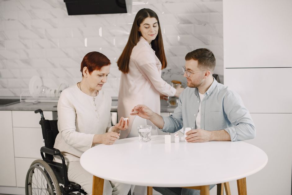 A man hands medication to a woman in a wheelchair at a table, while another woman—perhaps one of the family caregivers—stands by the kitchen counter in the background, highlighting the importance of respite care and support for those providing daily assistance.