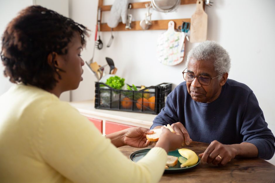 An elderly man sits at a table with food while a younger woman, part of a meal preparation service in Brazoria County, hands him a sandwich in a cozy kitchen setting.