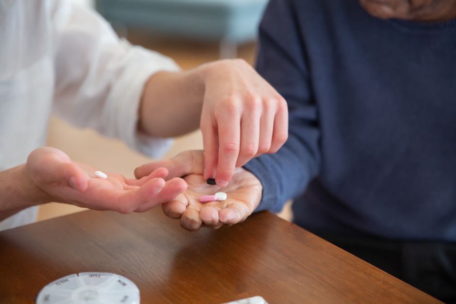 Two people sit at a table as one person places pills into the open palm of another, with a pill organizer nearby—a compassionate moment reflecting attentive Senior Care in a comfortable home setting.