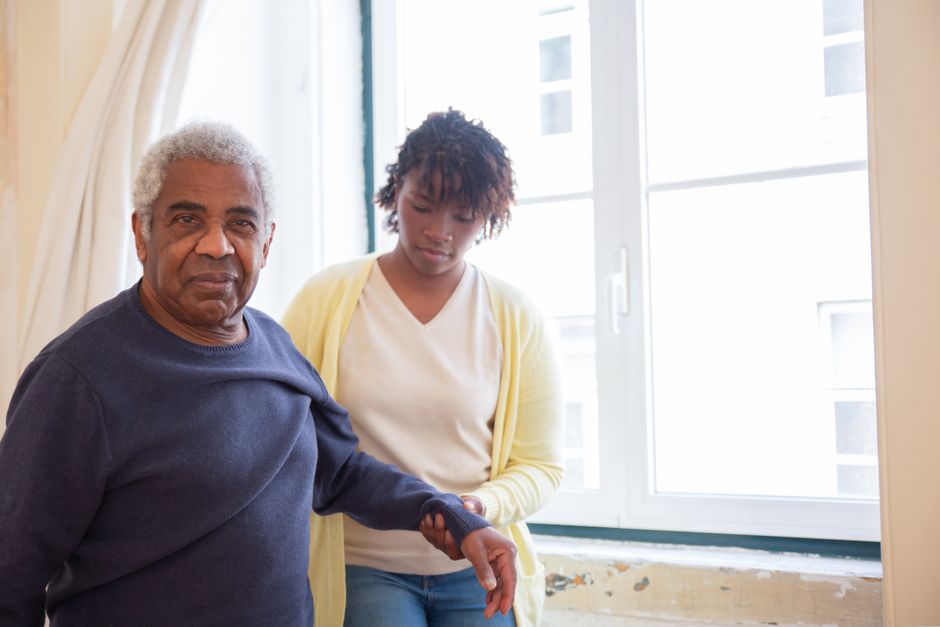 An elderly man stands holding hands with a younger woman near a window in a bright room, showcasing the warmth and support offered through Senior Companion Care in Lake Jackson.