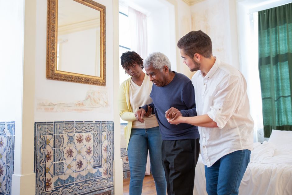 Two caregivers assist an elderly man walking in a room with patterned tiles, a large mirror, and a bed by the window—highlighting modern senior care options that support working families seeking peace of mind.