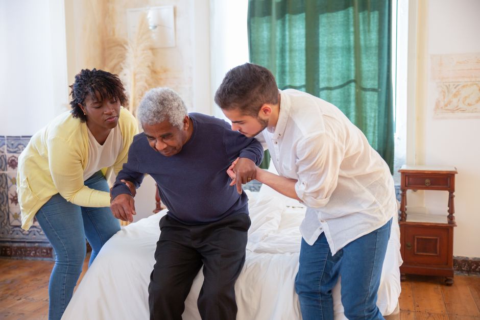 Two caregivers assist an elderly man as he stands up from a bed in a well-lit room with wooden floors and a green curtain, providing compassionate home assistance that supports aging in place for families seeking comfort and independence for their loved ones.
