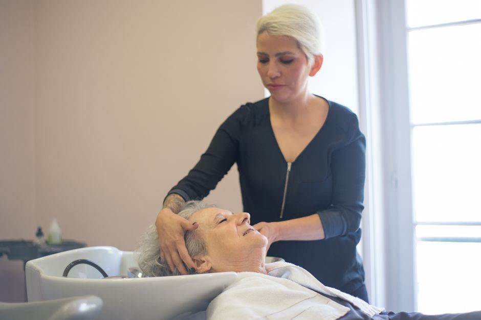 A hairdresser with short blonde hair washes an elderly woman’s hair at a salon sink, providing attentive Elderly Personal Care Services in Clute.