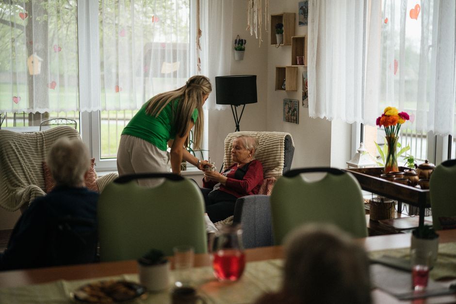 A caregiver provides in-home care in Alvin, TX, as she stands beside an elderly woman in an armchair and hands her a small object.