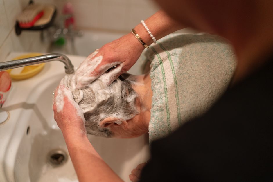 A caregiver provides elderly bathing assistance by washing the gray hair of someone leaning over a bathroom sink.