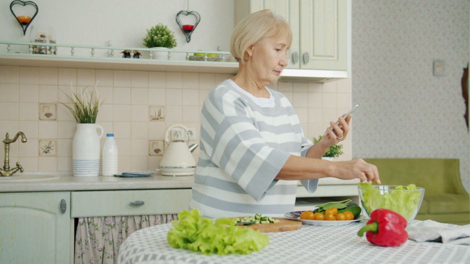 An older woman in a striped sweater uses her smartphone while preparing a salad in her kitchen, exemplifying senior independence.