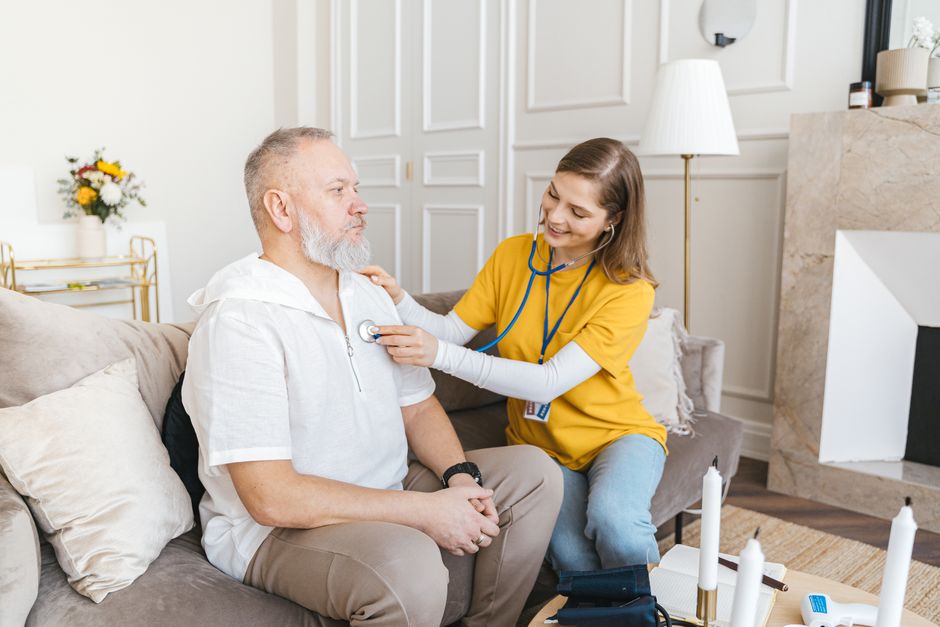 A healthcare worker uses a stethoscope to examine an older man receiving in-home care on a sofa.