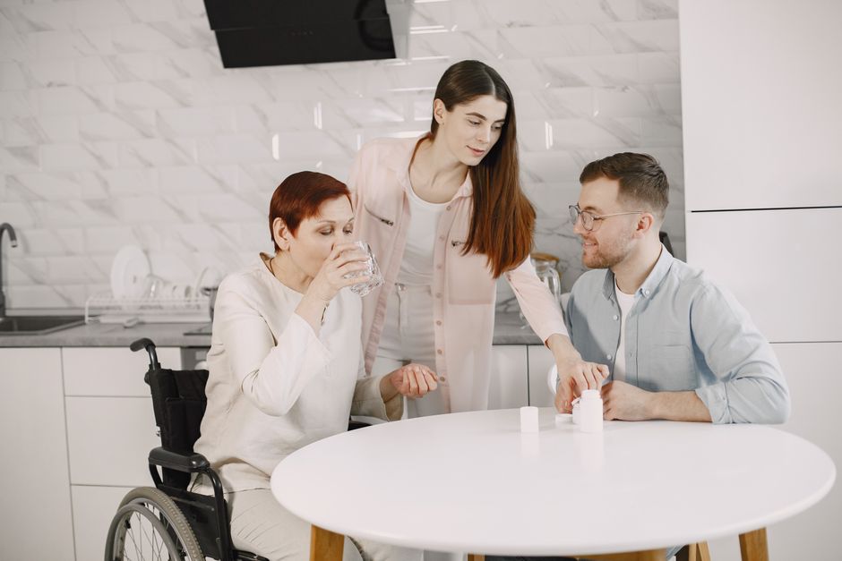 A young woman and man helping a woman in a wheelchair take her medication at a kitchen table, illustrating elderly assistance in Texas.
