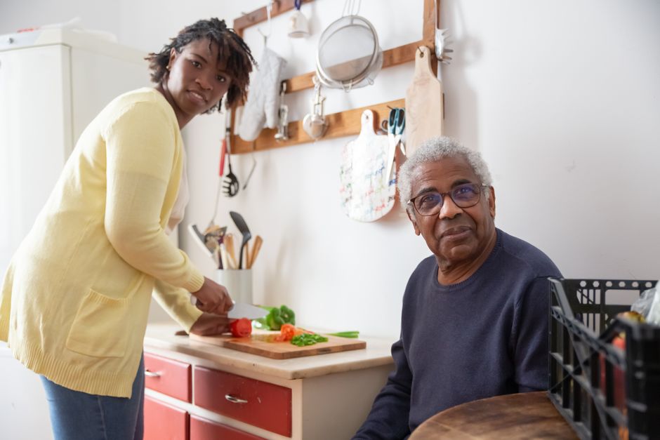 A caregiver guides a woman chopping vegetables on a kitchen counter while an older man sits nearby.