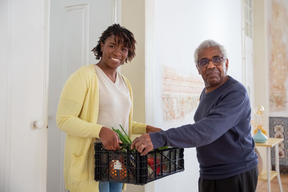 A young woman and an older man hold a plastic crate filled with fresh vegetables, part of a home organization guide for seniors.
