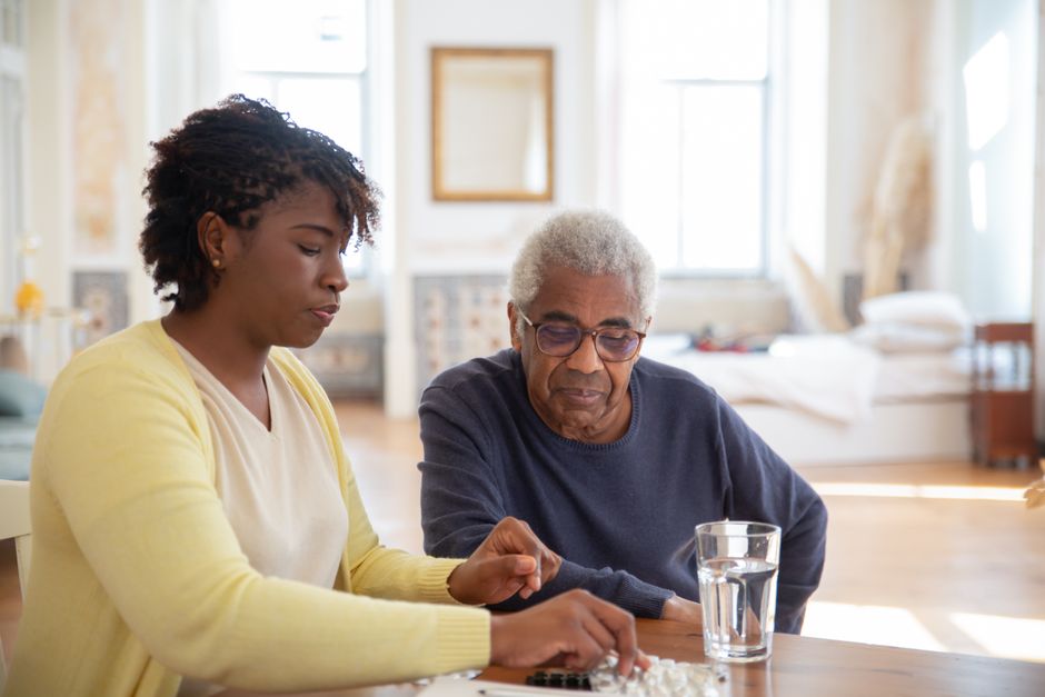 A woman in a yellow cardigan helps an older man organize medication from a pill organizer on a wooden table, providing professional in-home care.