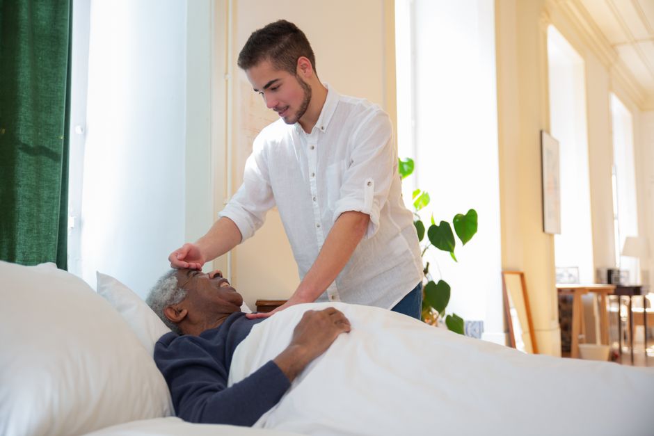 A caregiver in a white shirt gently touches the forehead of an elderly man lying in a bed, illustrating respectful elder care.