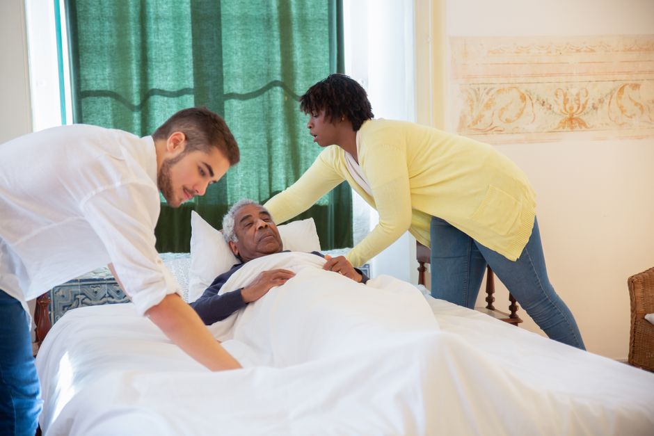 Two caregivers adjust the bedding for an elderly man lying in a hospital bed as part of our senior home care services in Pearland, TX.