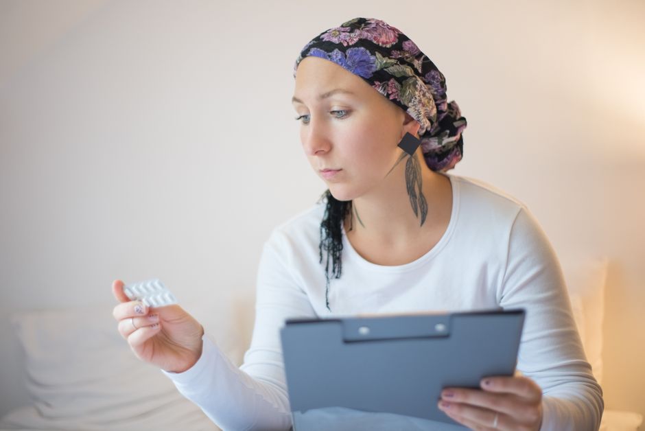 An elderly woman wearing a floral headscarf holds a clipboard and reviews a blister pack of pills as part of her cancer recovery.