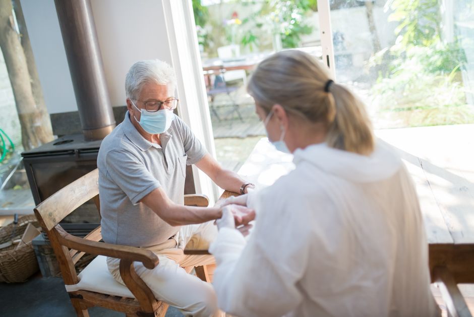 An older man and a woman, both wearing face masks, sit across from each other at a wooden table—offering support and preventing caregiver burnout in a well-lit room with large windows.