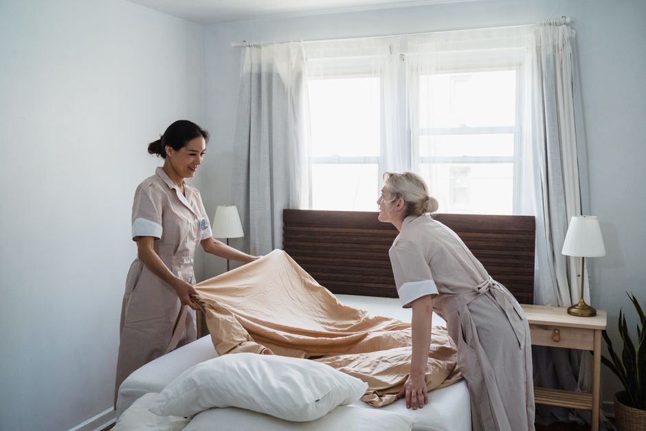 Two housekeepers in uniforms providing light housekeeping for seniors as they make a hotel bed.